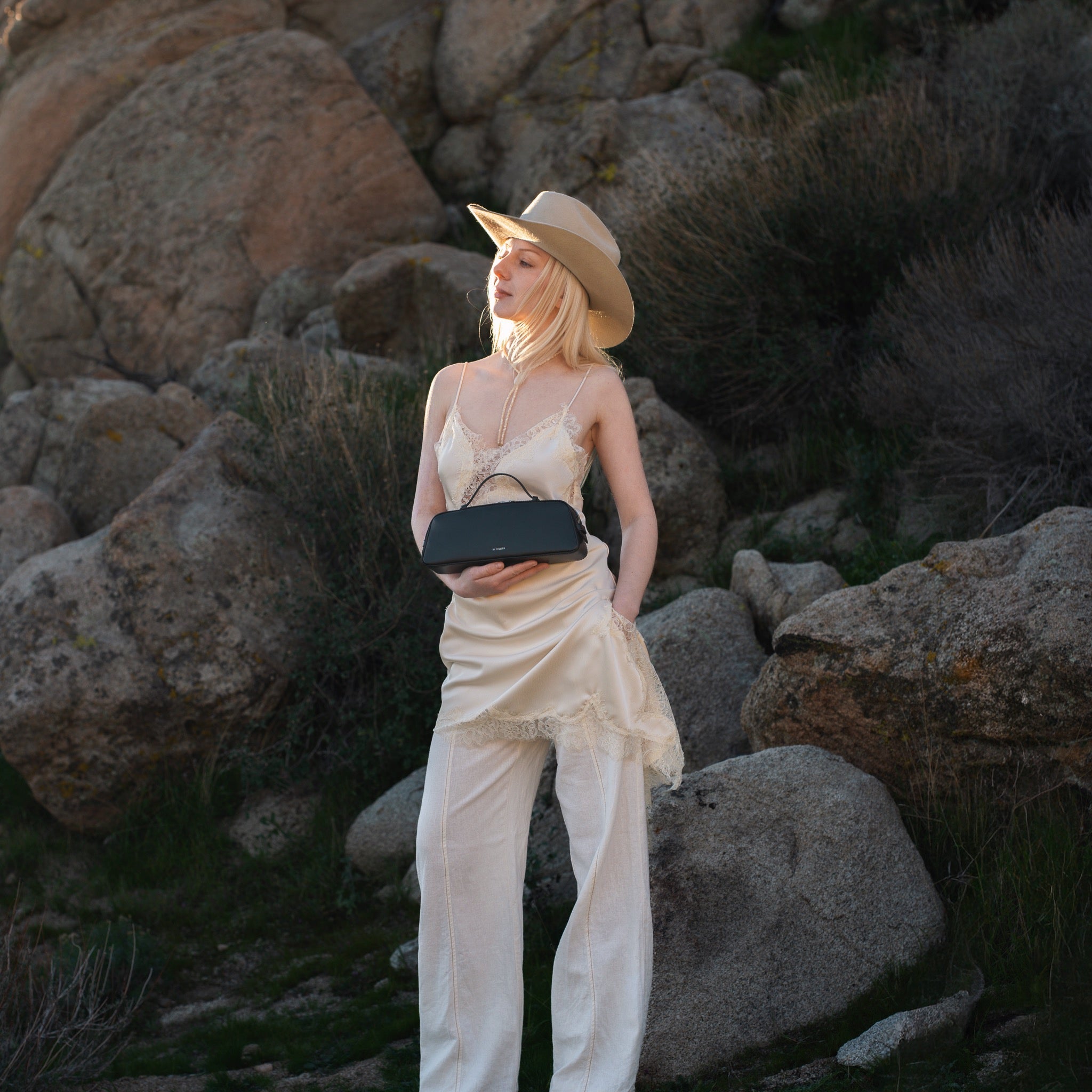 Woman in a white dress and hat holding a black clutch against a rocky background