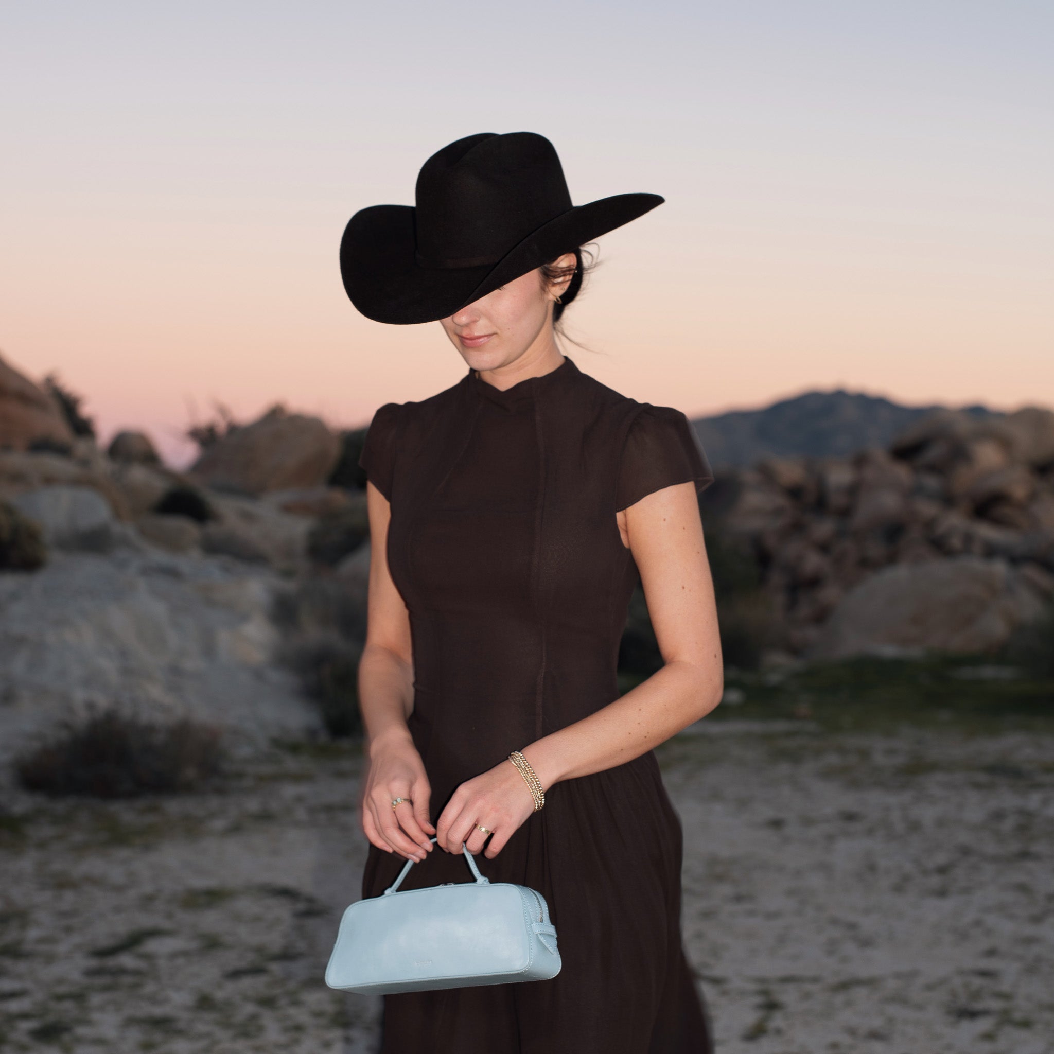 Woman in a dark dress and black hat holding a light blue handbag in a desert landscape.