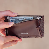 Brown wallet with money held open by a hand on a beige background, brand 'BY COLLIER' visible.